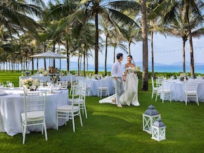 A wedding reception with tables set outdoors, surrounded by palm trees and a view of the sea.