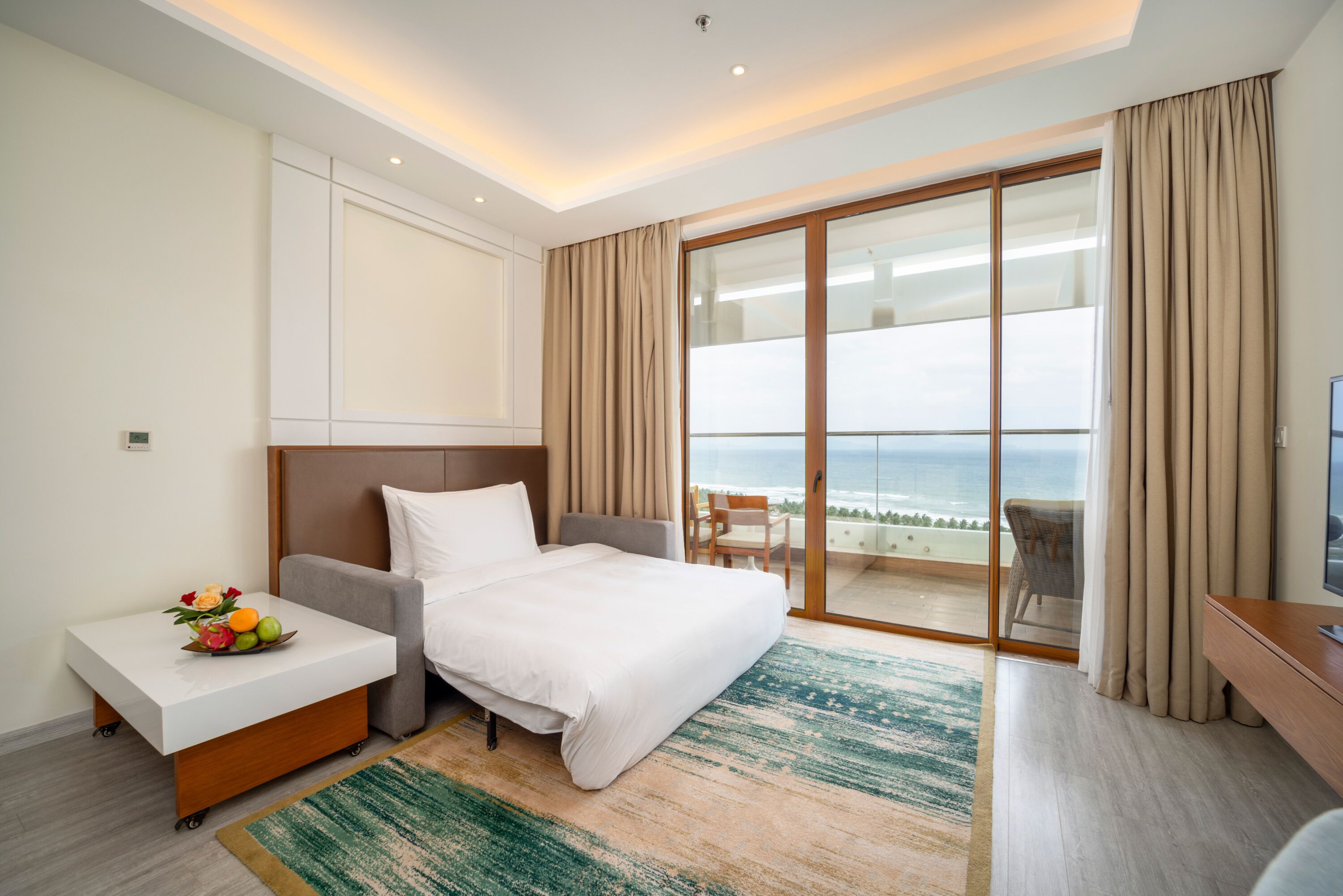 Habitación de hotel con cama, una mesita con fruta, televisor y balcón con vista al océano.