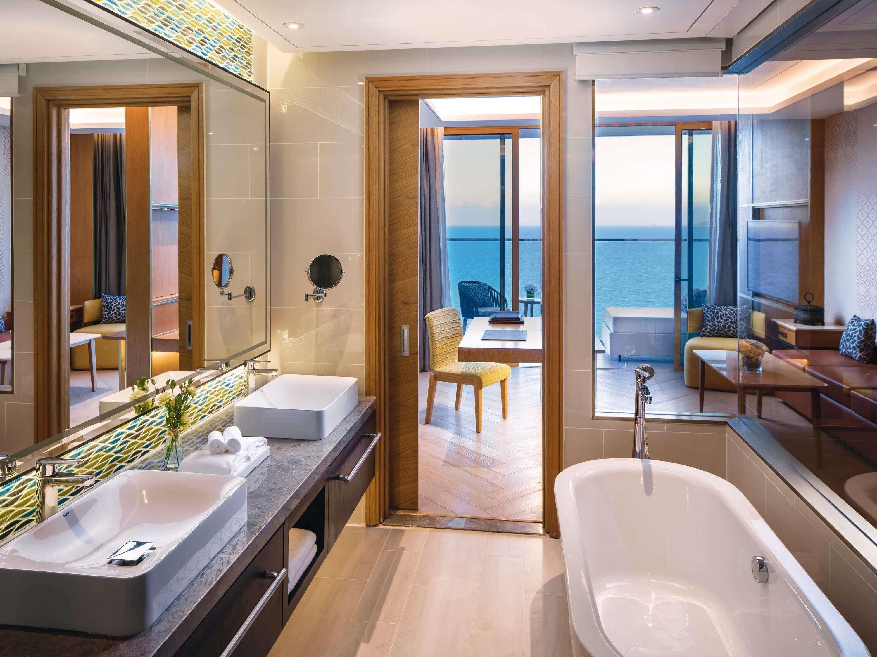 A modern bathroom with a large bathtub, a vanity with two sinks, and a large mirror. There is a view of the ocean through large windows.