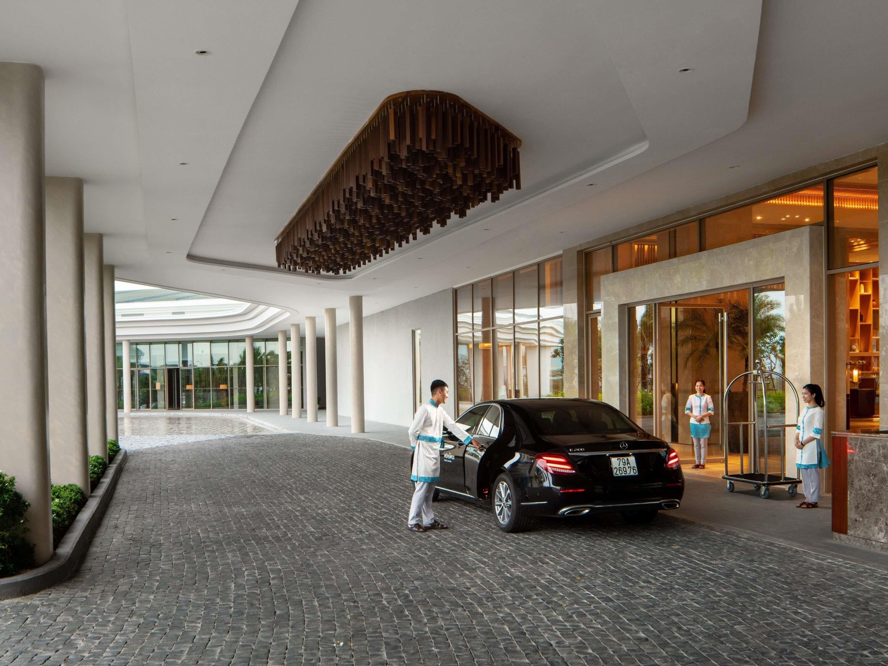 A modern hotel entrance with a black car parked in front, a person interacting with the car, and a large glass door leading inside.