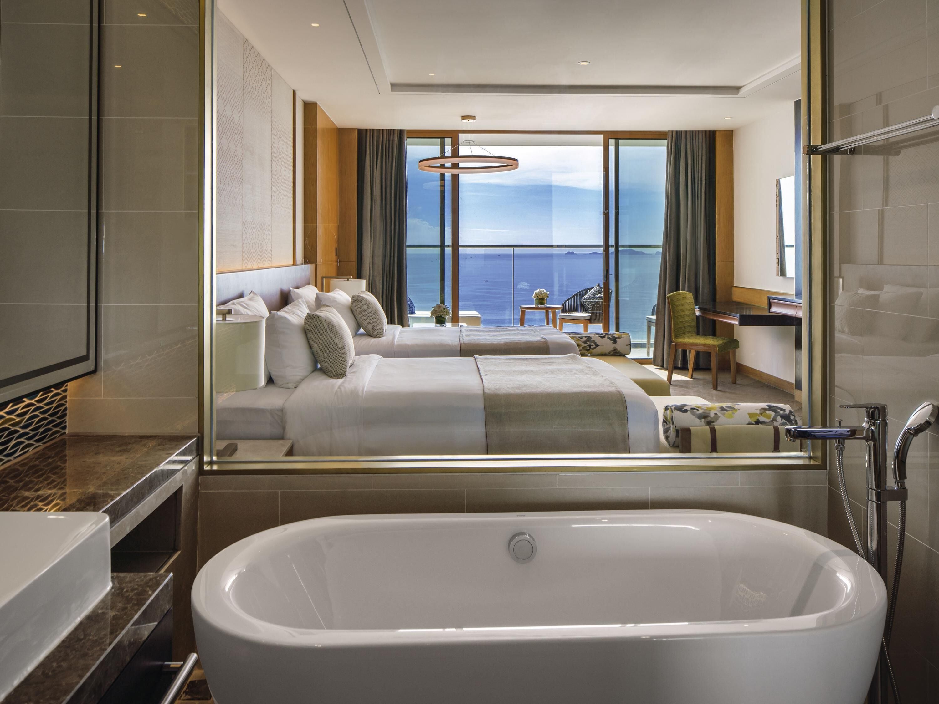 Habitación de hotel con una bañera grande, dos camas, un área de descanso con vistas al mar y un espejo grande que refleja la habitación.