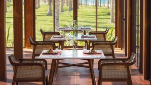 An outdoor dining area with tables and chairs, a view of a grassy landscape and palm trees, and a wooden pergola structure.