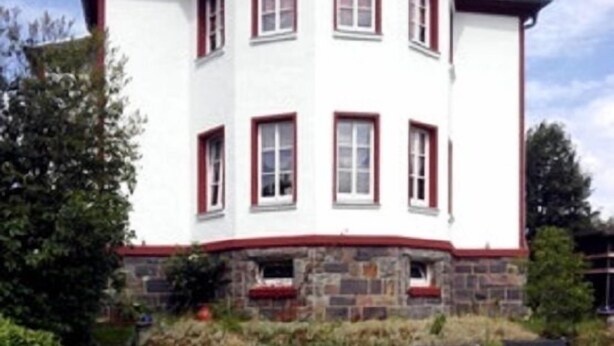 A white building with red trim and stone foundation. There are four black lounge chairs in front. - Ferienhaus XXL Deutschland (Schwarzatal)