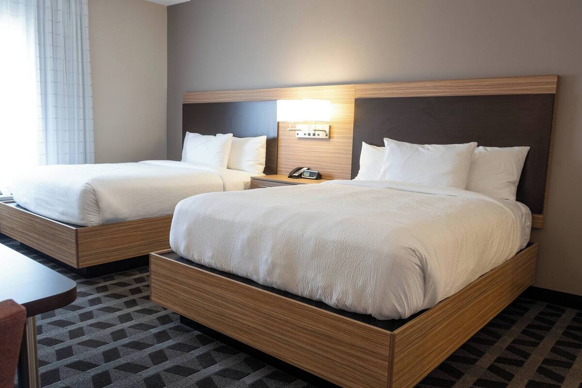 Habitación de hotel con dos camas individuales, una mesita de noche con lámpara y un escritorio pequeño con silla.
