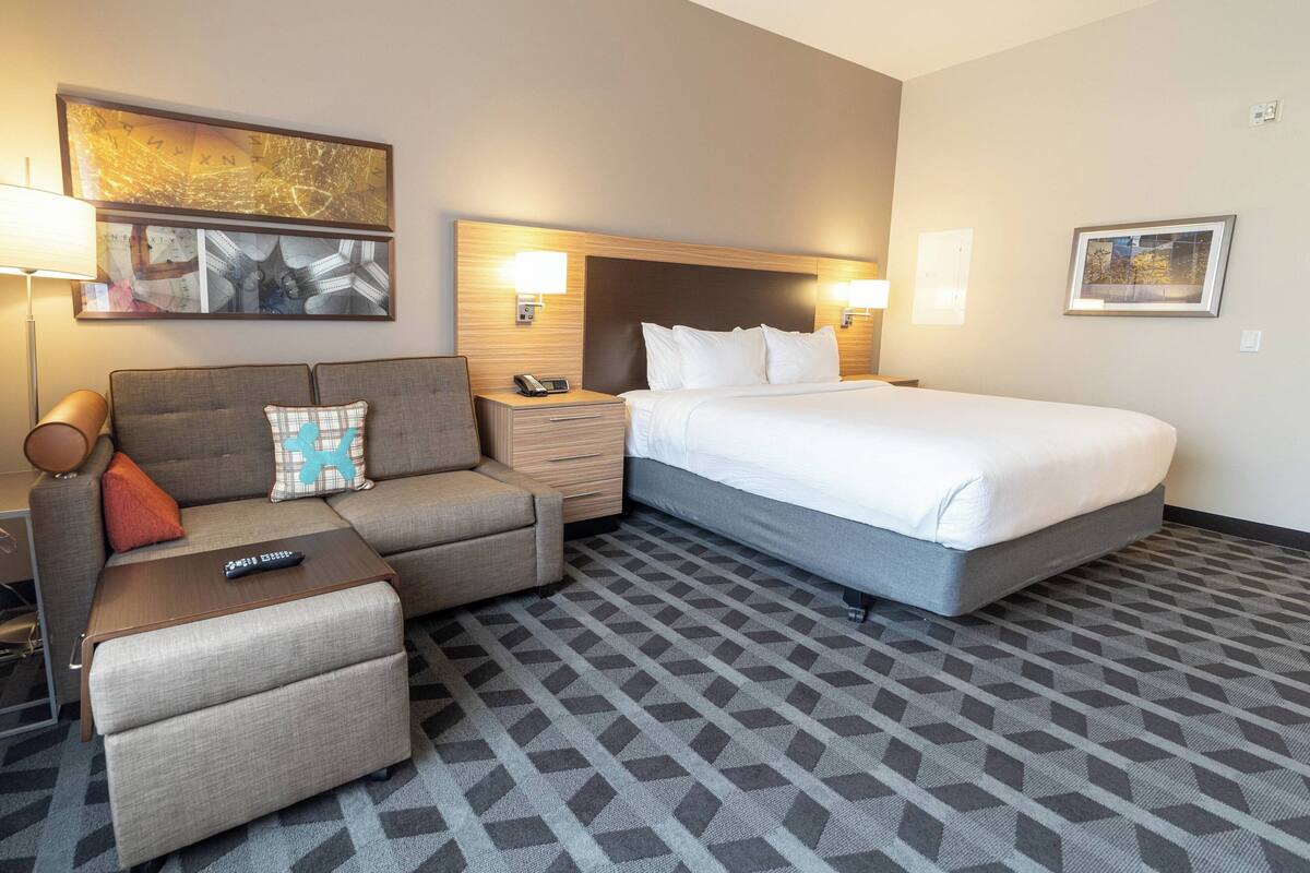 Habitación de hotel con cama, sofá, mesita pequeña, mesita de noche y lámpara.
