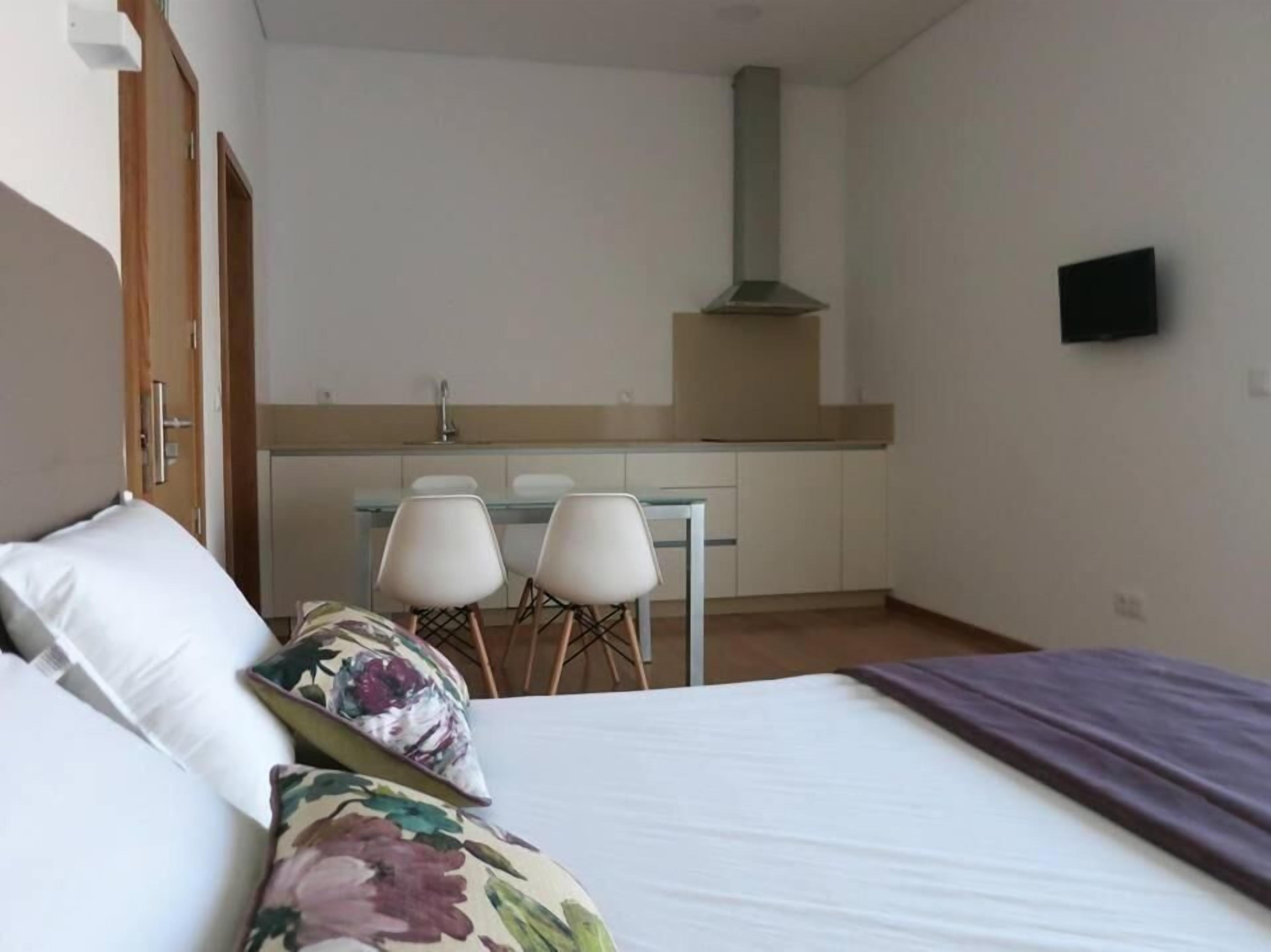 A bedroom with a bed, two chairs, a small kitchenette, and a TV mounted on the wall.