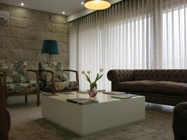 Una sala de estar con pared de piedra, un sofá marrón, dos sillones con motivos florales, una mesa de centro con libros y un jarrón de flores, y una lámpara azul.