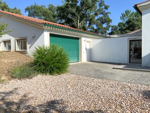 A white building with a green garage door, a gravel driveway, and a small bush in the foreground.