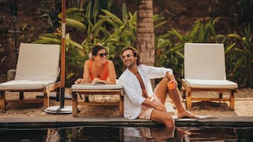 Two people sitting by a poolside lounge chair, with a palm tree and greenery in the background.