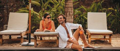 Two people sitting by a poolside lounge chair, with a palm tree and greenery in the background.