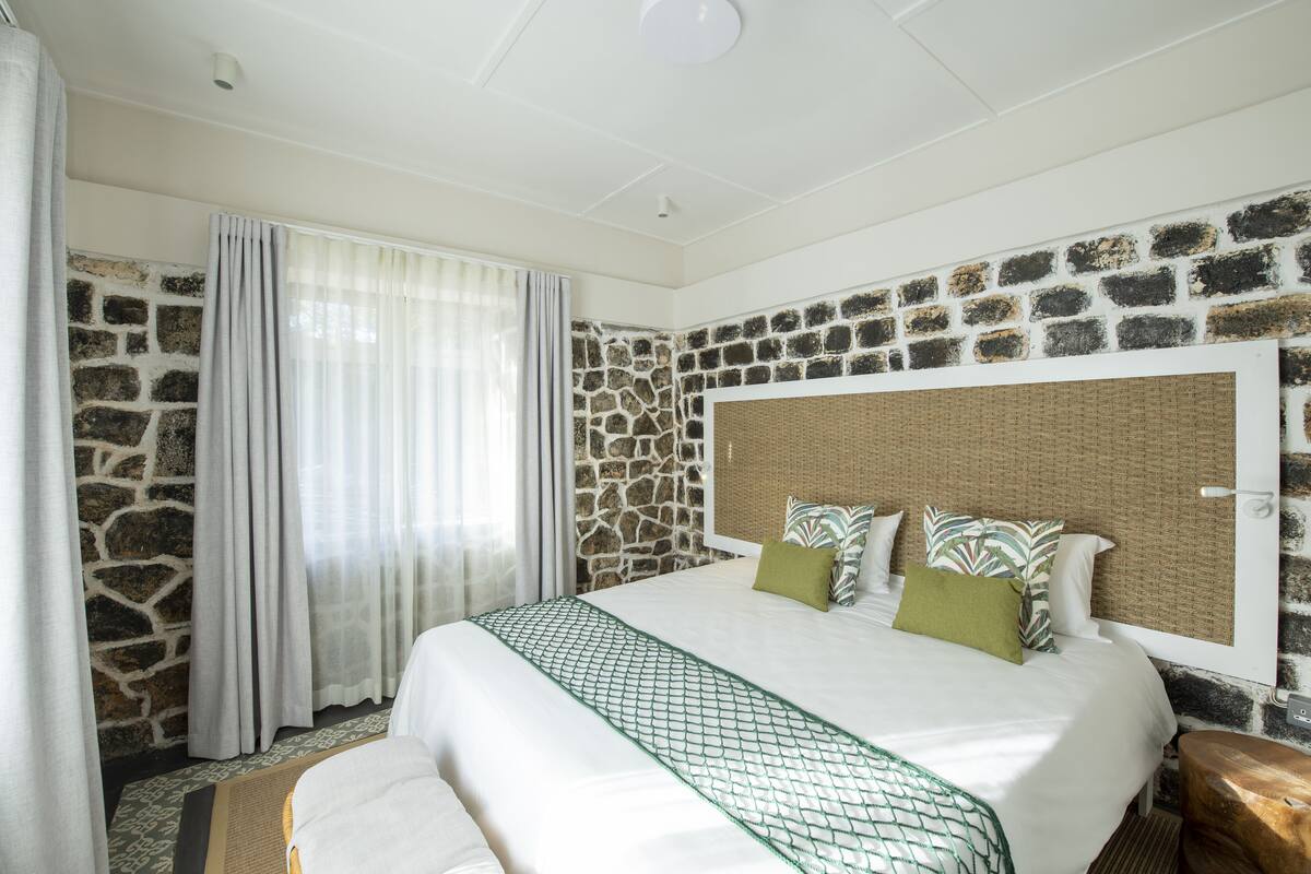 A hotel room with a stone wall, a bed with a green netted throw, and a wooden nightstand.