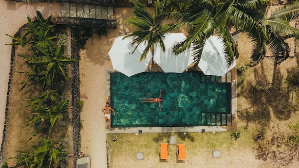 Vista aérea de una piscina con una persona nadando, rodeada de palmeras y una zona arenosa con dos sillas de playa.