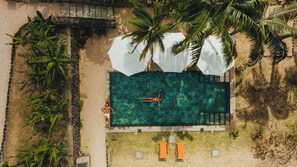 Vista aérea de una piscina con una persona nadando, rodeada de palmeras y una zona arenosa con dos sillas de playa.