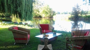 An outdoor seating area with wicker furniture and a table, surrounded by a grassy lawn and a body of water.