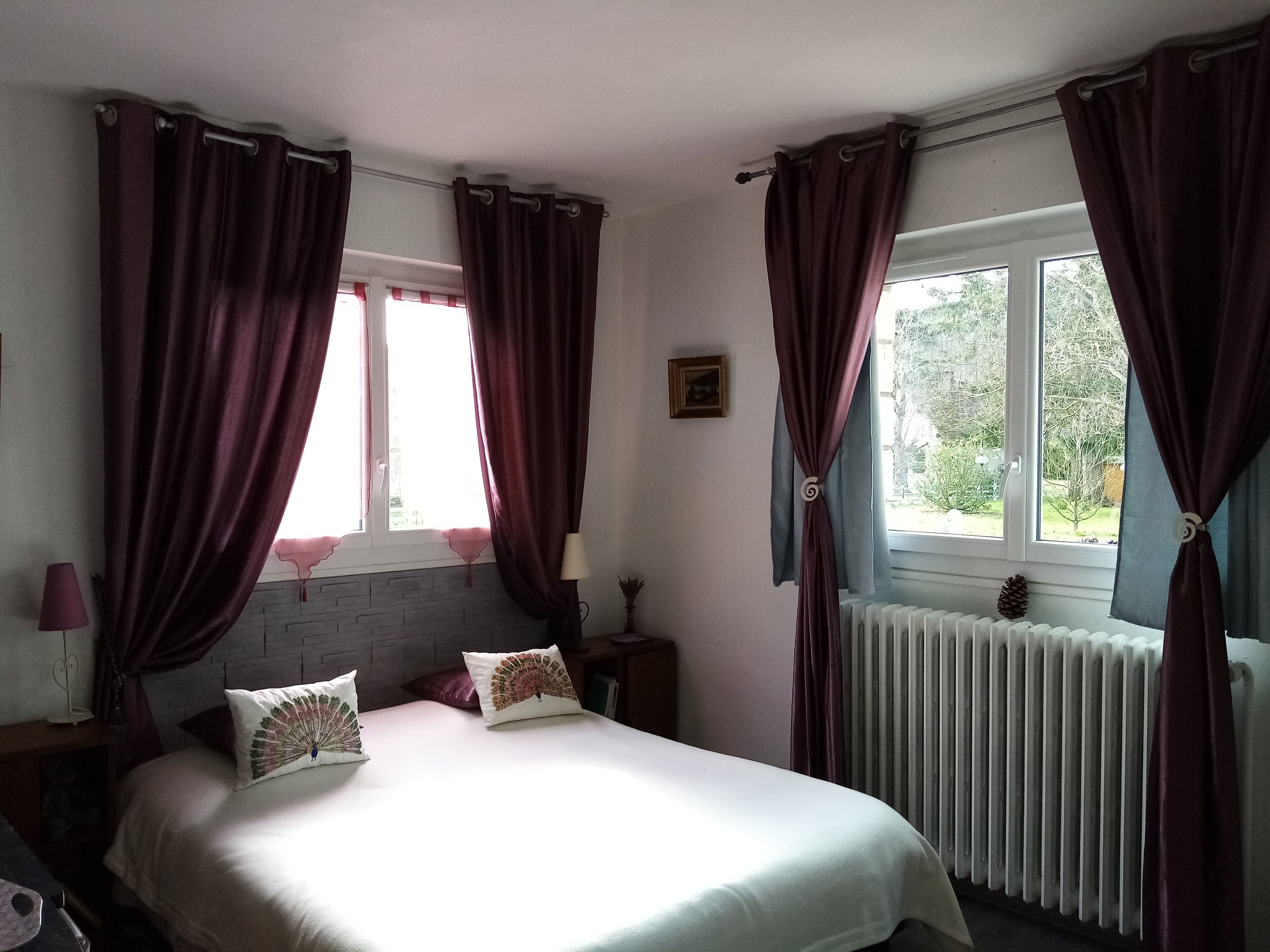 Un dormitorio con una cama, dos almohadas, una mesita de noche, una lámpara, una ventana con cortinas y un radiador.