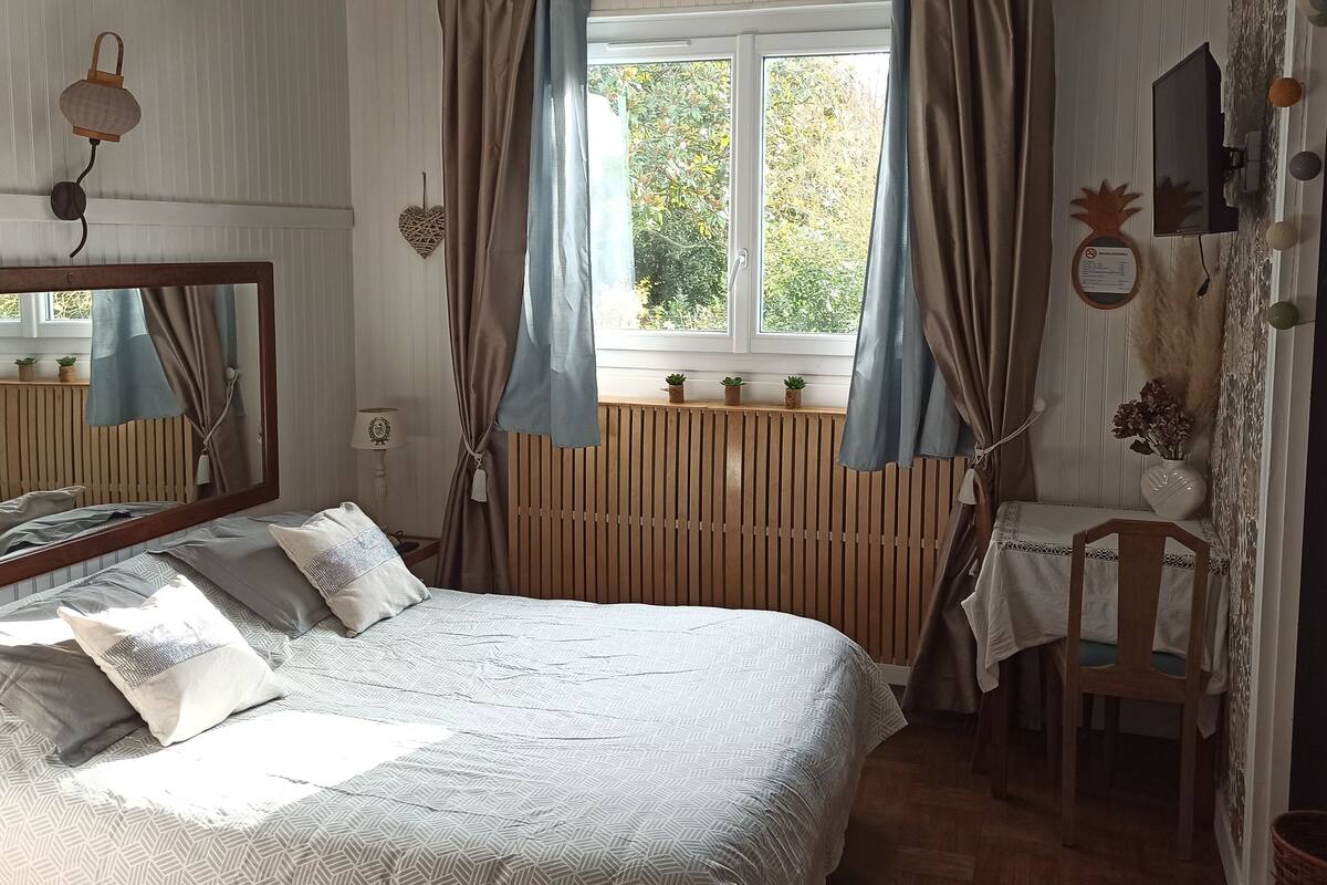 A bedroom with a bed, a desk, a chair, a window with curtains, a mirror, and a wall-mounted TV.