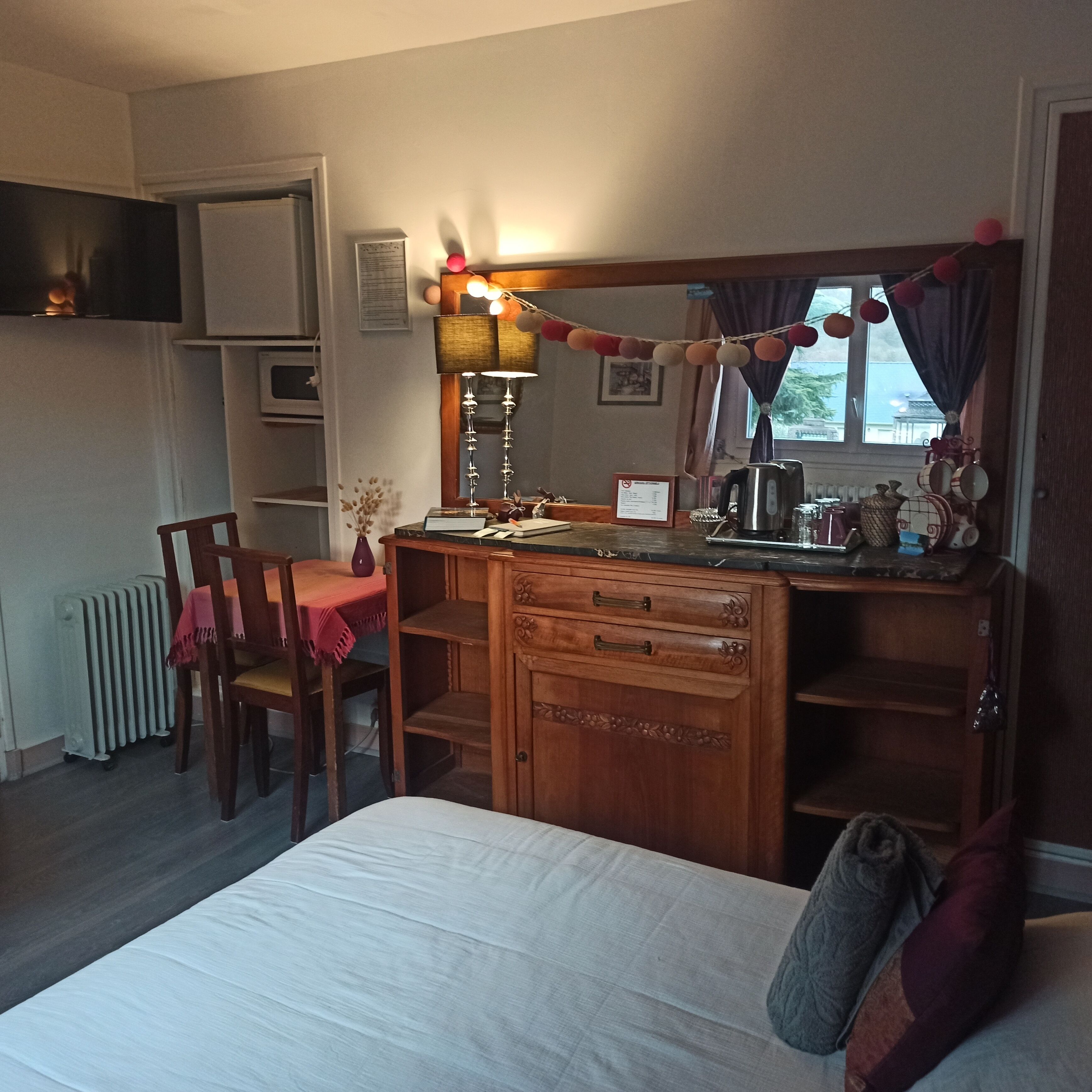 A bedroom with a wooden dresser, a bed, a desk with a lamp, a radiator, and a window with curtains.