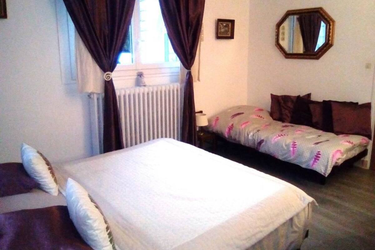A bedroom with a bed, a sofa, a window with curtains, a radiator, and a mirror on the wall.