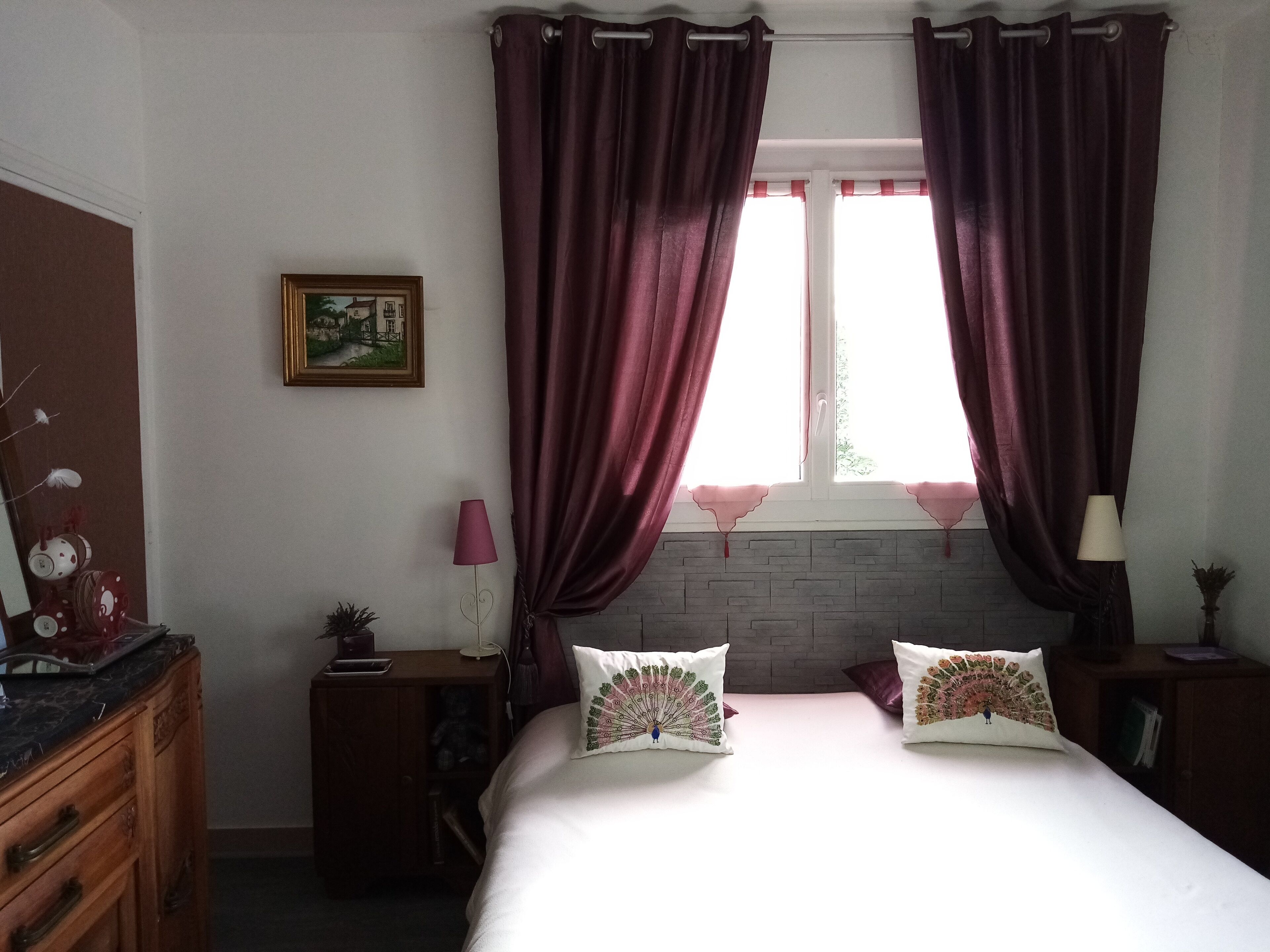 Un dormitorio con cama, dos almohadas, una mesita de noche, una lámpara, un aparador, una ventana con cortinas y un cuadro en la pared.