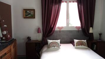 Un dormitorio con cama, dos almohadas, una mesita de noche, una lámpara, un aparador, una ventana con cortinas y un cuadro en la pared.