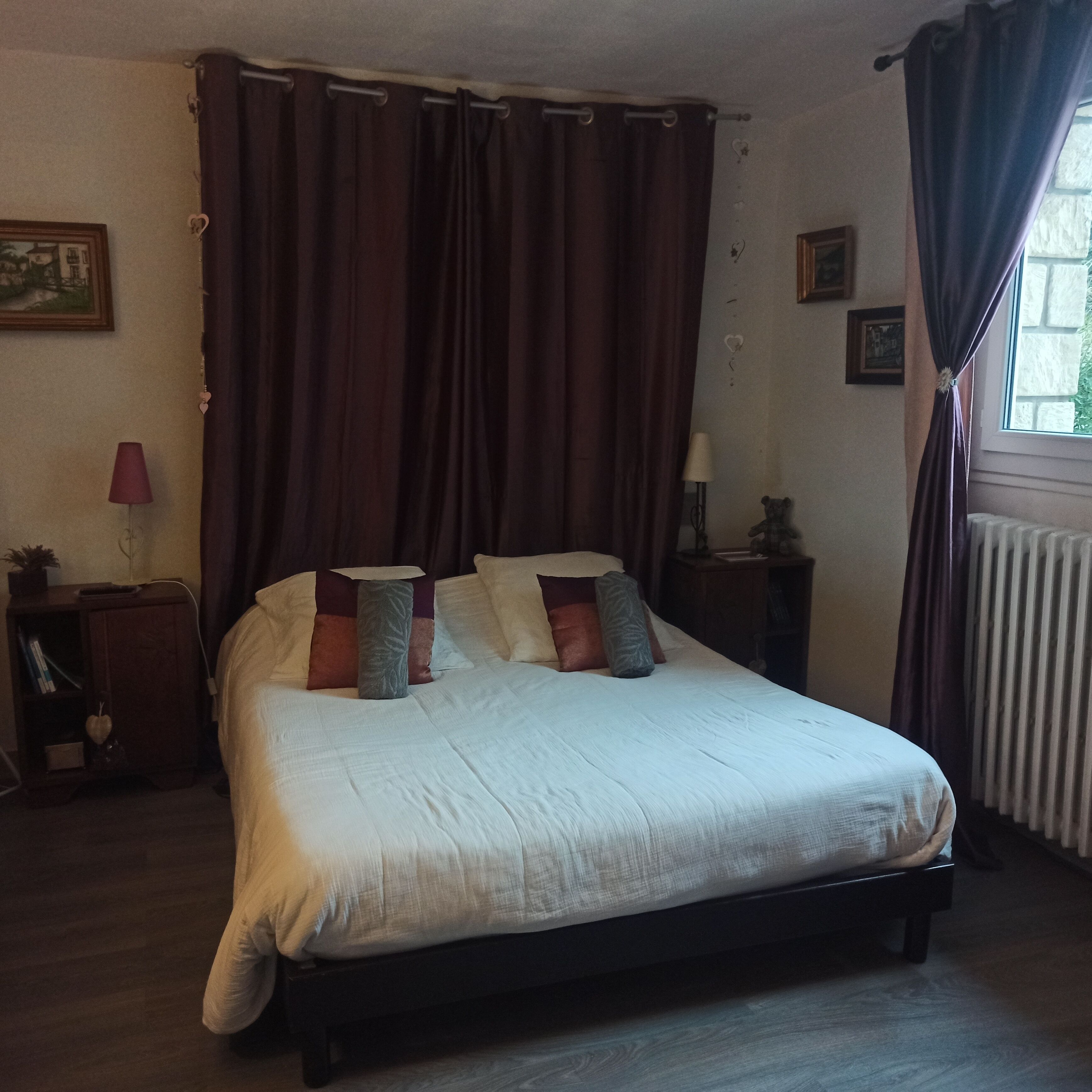 Un dormitorio con una cama grande, cortinas oscuras, una mesita de noche con lámpara y un radiador.
