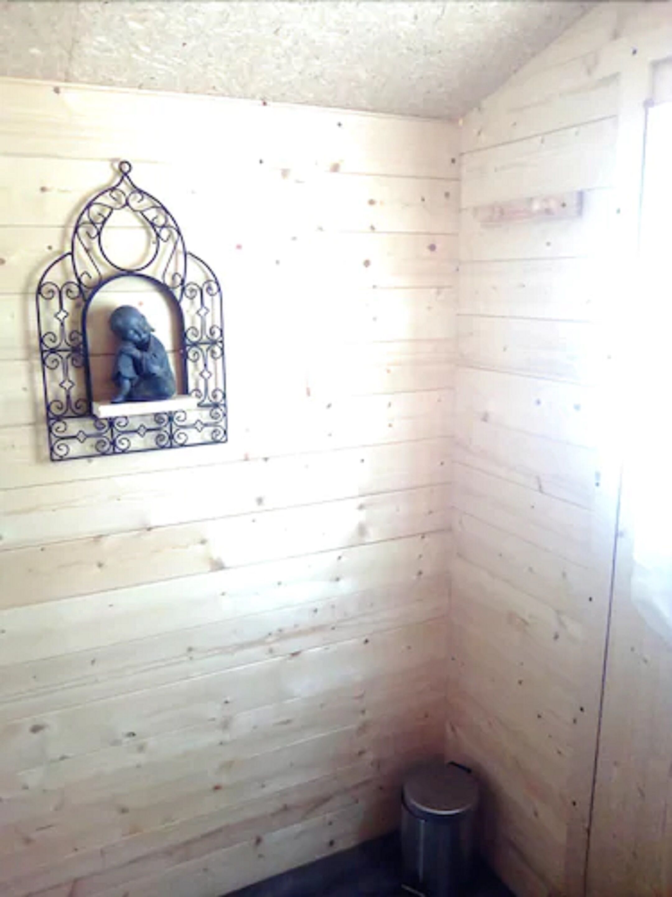 A corner of a room with wooden walls, a small trash bin, and a decorative metal mirror with a sculpture.