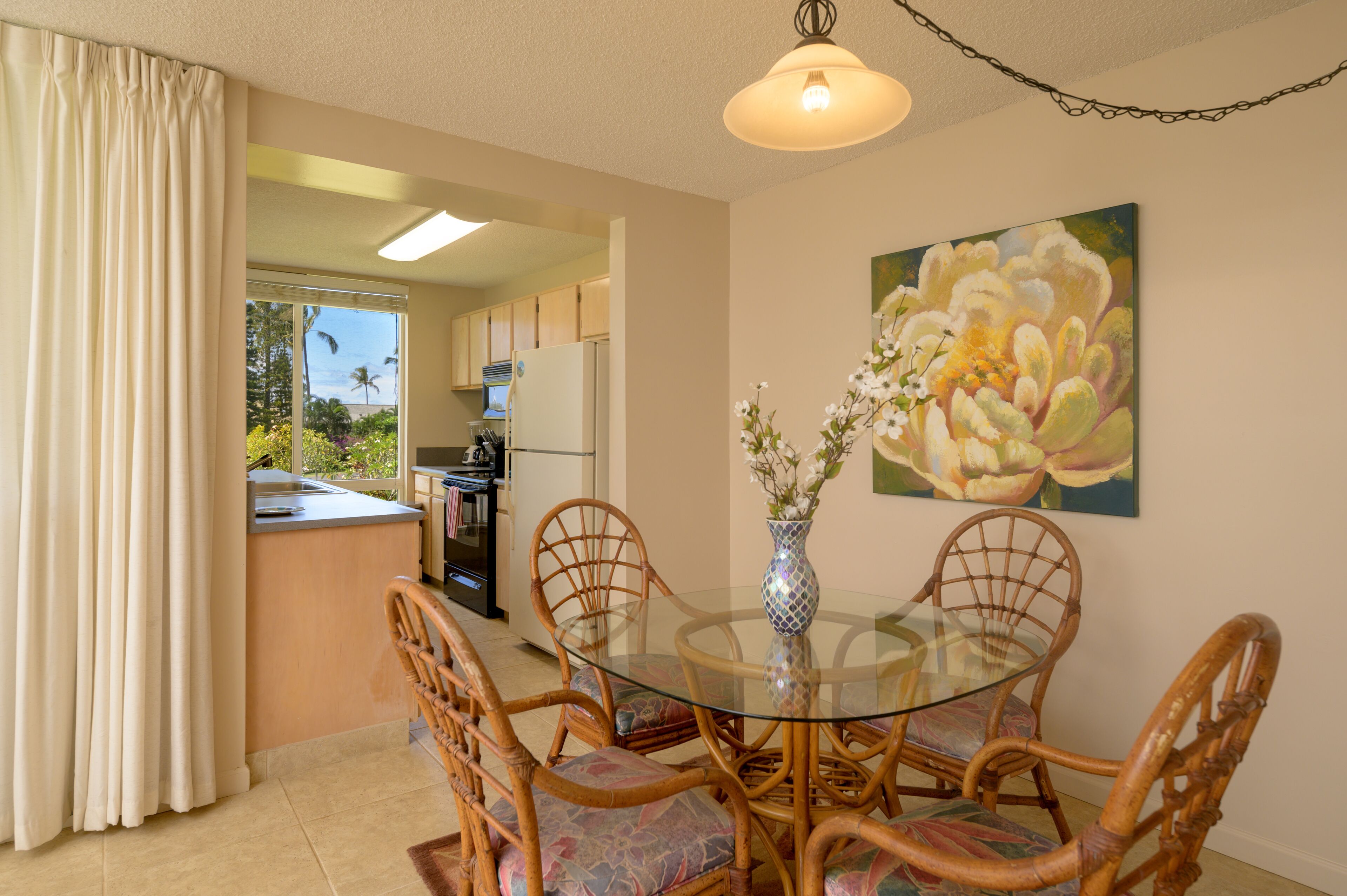 A dining area with a glass table surrounded by wicker chairs, a vase with flowers, and a painting of flowers on the wall.