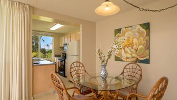 A dining area with a glass table surrounded by wicker chairs, a vase with flowers, and a painting of flowers on the wall.