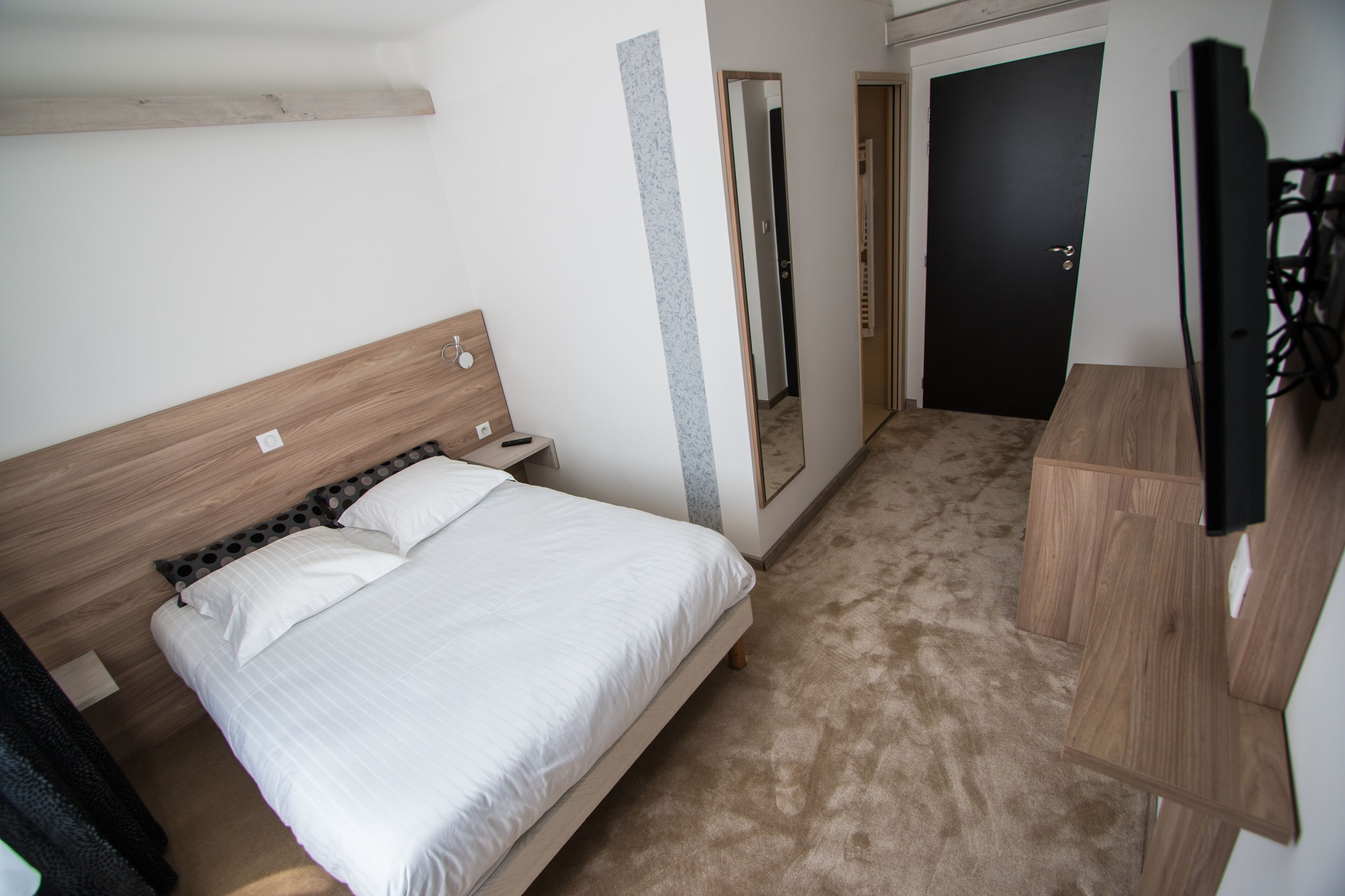 Un dormitorio con cama de madera, mesita de noche, televisor montado en la pared y puerta que da a otra habitación.
