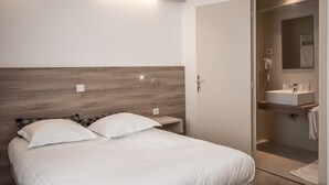 Una habitación de hotel moderna con cabecera de madera, cama con ropa blanca, mesita de noche y baño con lavamanos y espejo.
