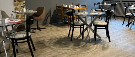 Un espace repas doté d’un plancher en bois, de tables et de chaises. Deux tableaux ornent le mur : l’un représente une carte du monde, l’autre une voiture sur un fond rouge.