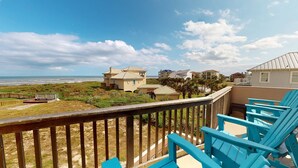 Une propriété située en bord de mer, dotée d’une terrasse en bois et de chaises bleues, offrant une vue sur l’océan et les maisons voisines.
