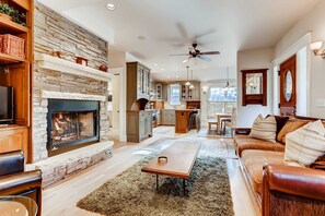 A spacious living area with a fireplace, a ceiling fan, and a kitchen in the background. - Historic High Street | Walk to Everything | Charm! (Breckenridge)