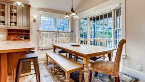 Zona pranzo con tavolo in legno, panche e sedie, cucina con armadietti e ampie finestre con vista.