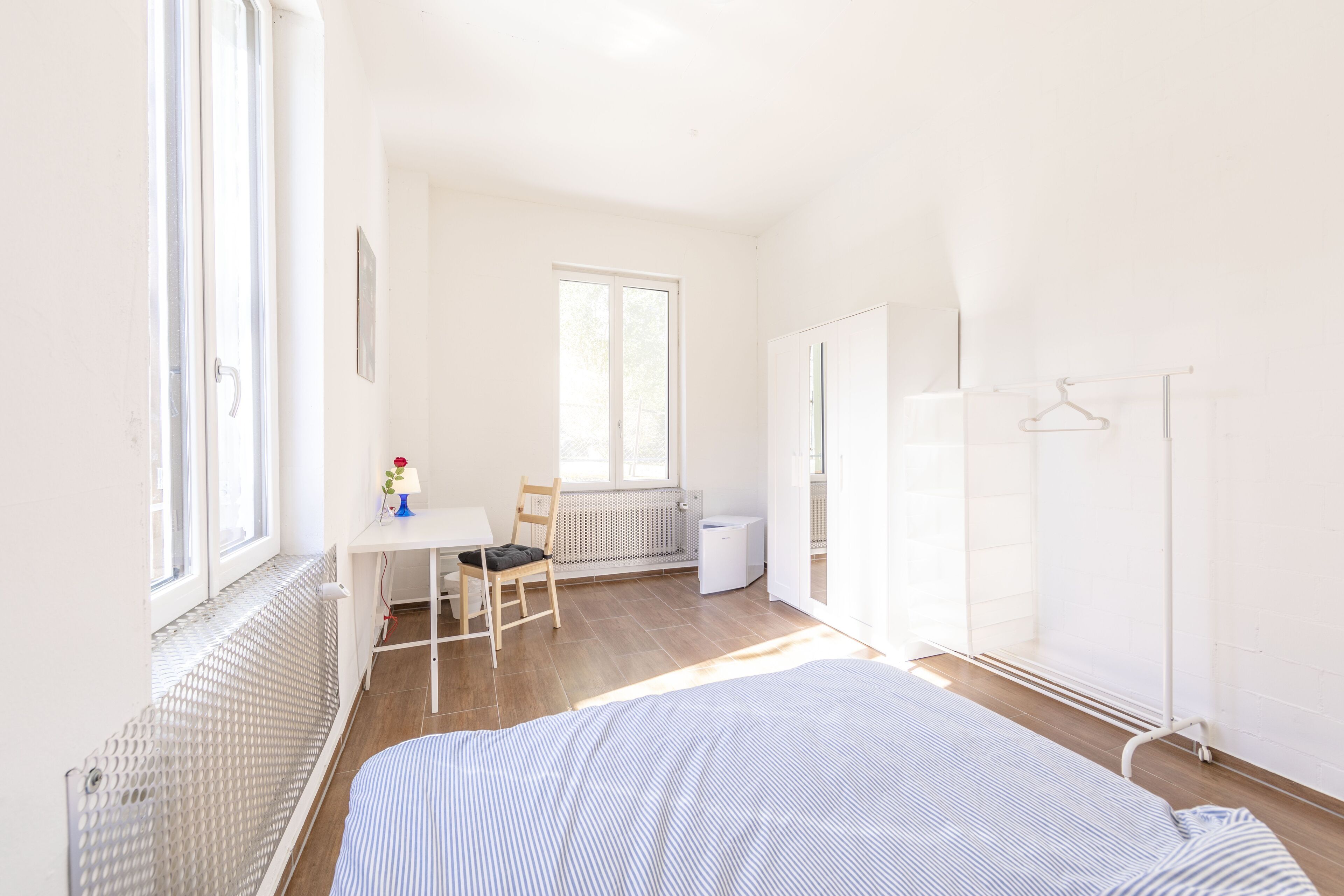 Ein heller, gut beleuchteter Raum mit einem Bett, einem Schreibtisch und einem Stuhl. Unter dem Schreibtisch befindet sich eine Heizung und ein Fenster sorgt für natürliches Licht.