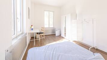 Ein heller, gut beleuchteter Raum mit einem Bett, einem Schreibtisch und einem Stuhl. Unter dem Schreibtisch befindet sich eine Heizung und ein Fenster sorgt für natürliches Licht.