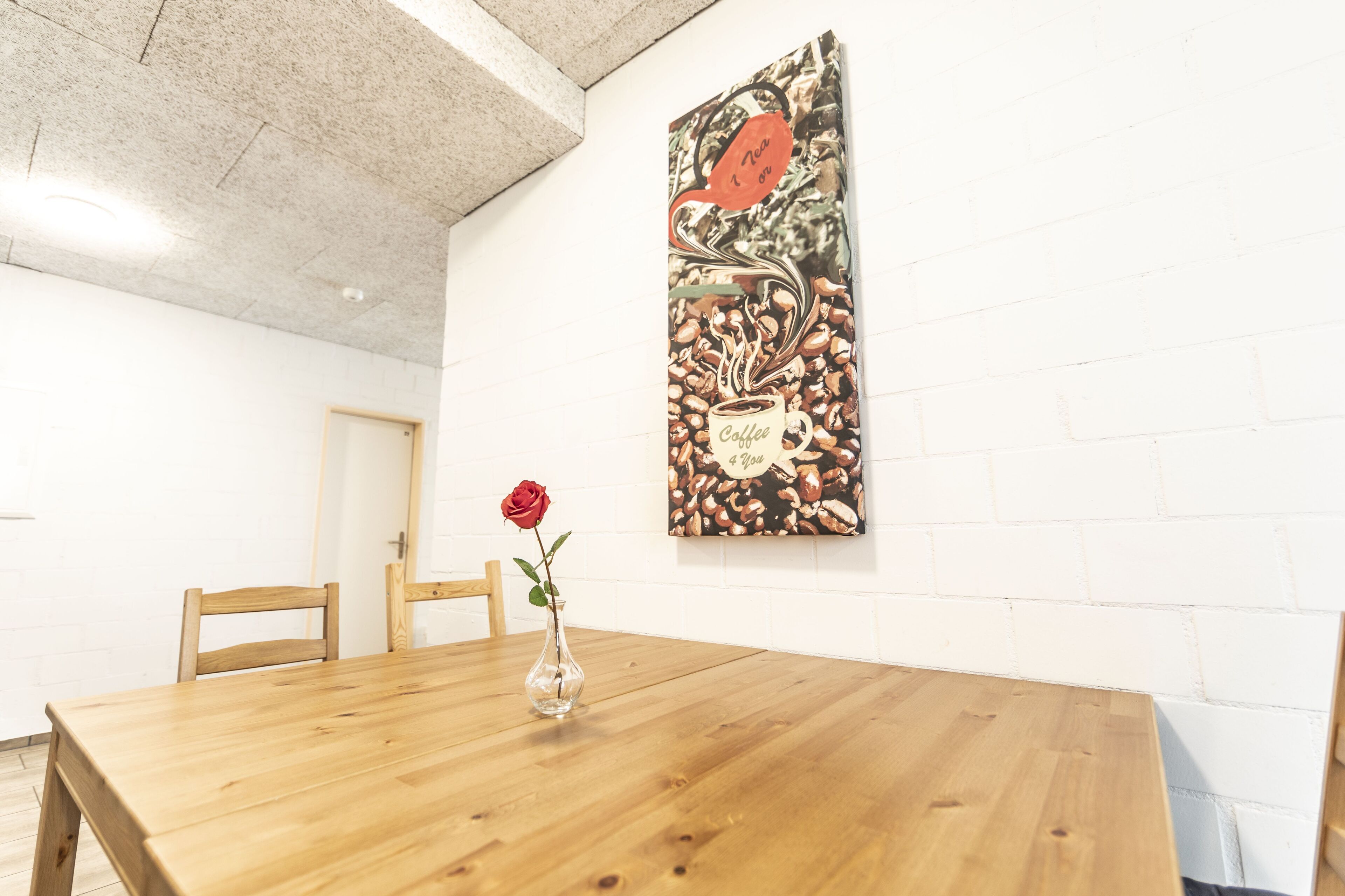 Una taula de fusta amb un ram de rosa vermella, una obra d'art al mur amb tema de cafè, i una porta al fons.