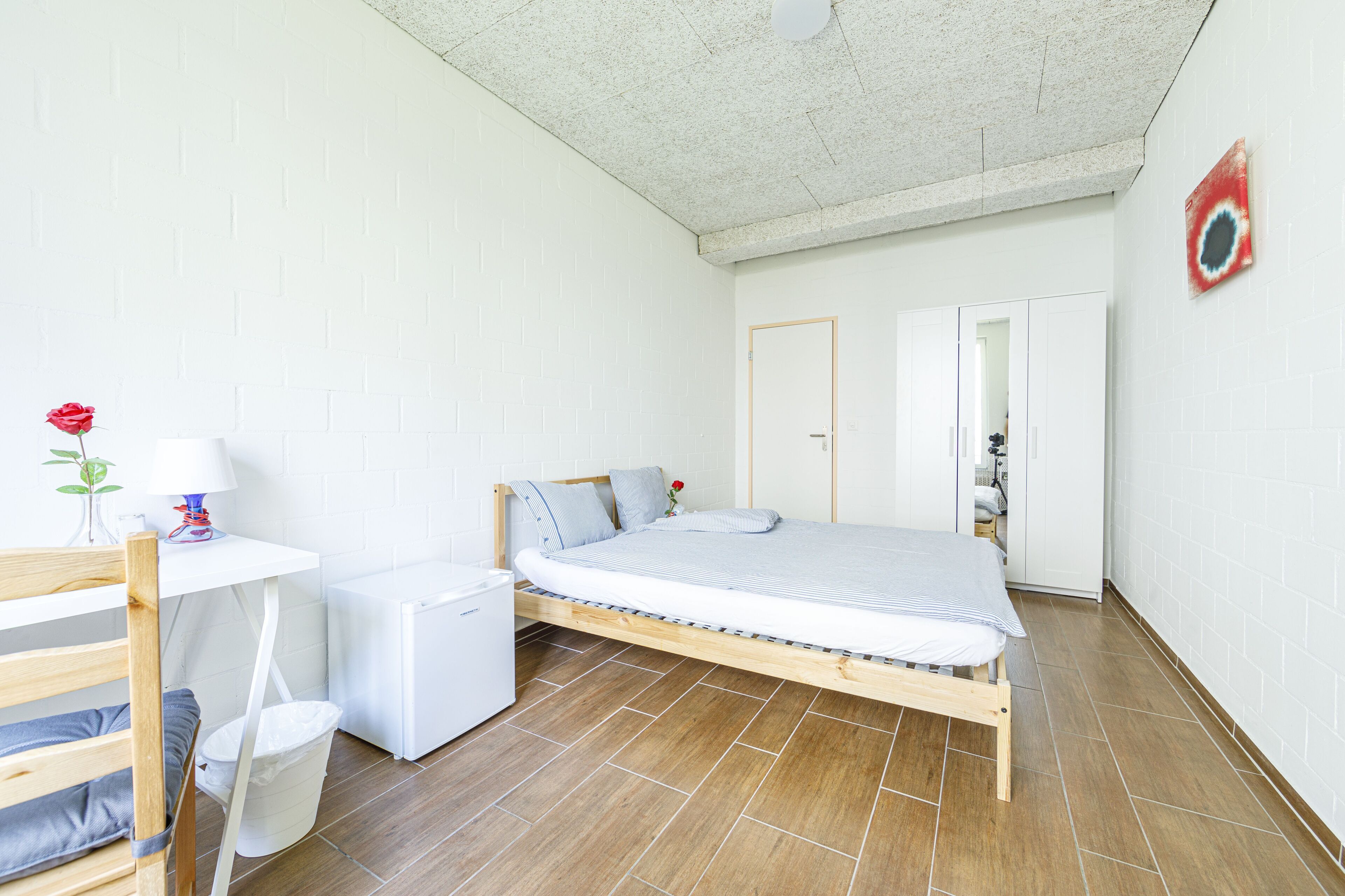 A compact bedroom with a single bed, a small table with a lamp, a chair, and a white radiator.