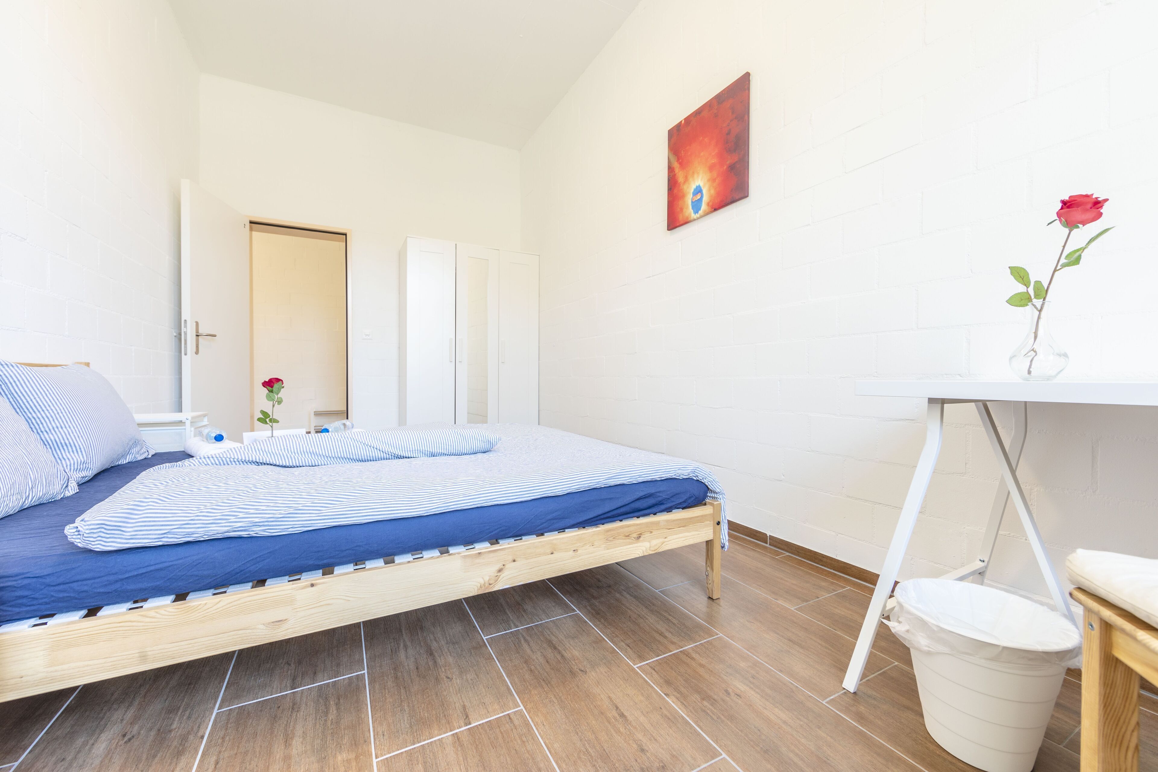 Une chambre à coucher avec un lit en bois, un bureau blanc, une poubelle blanche et une rose rouge dans un vase.