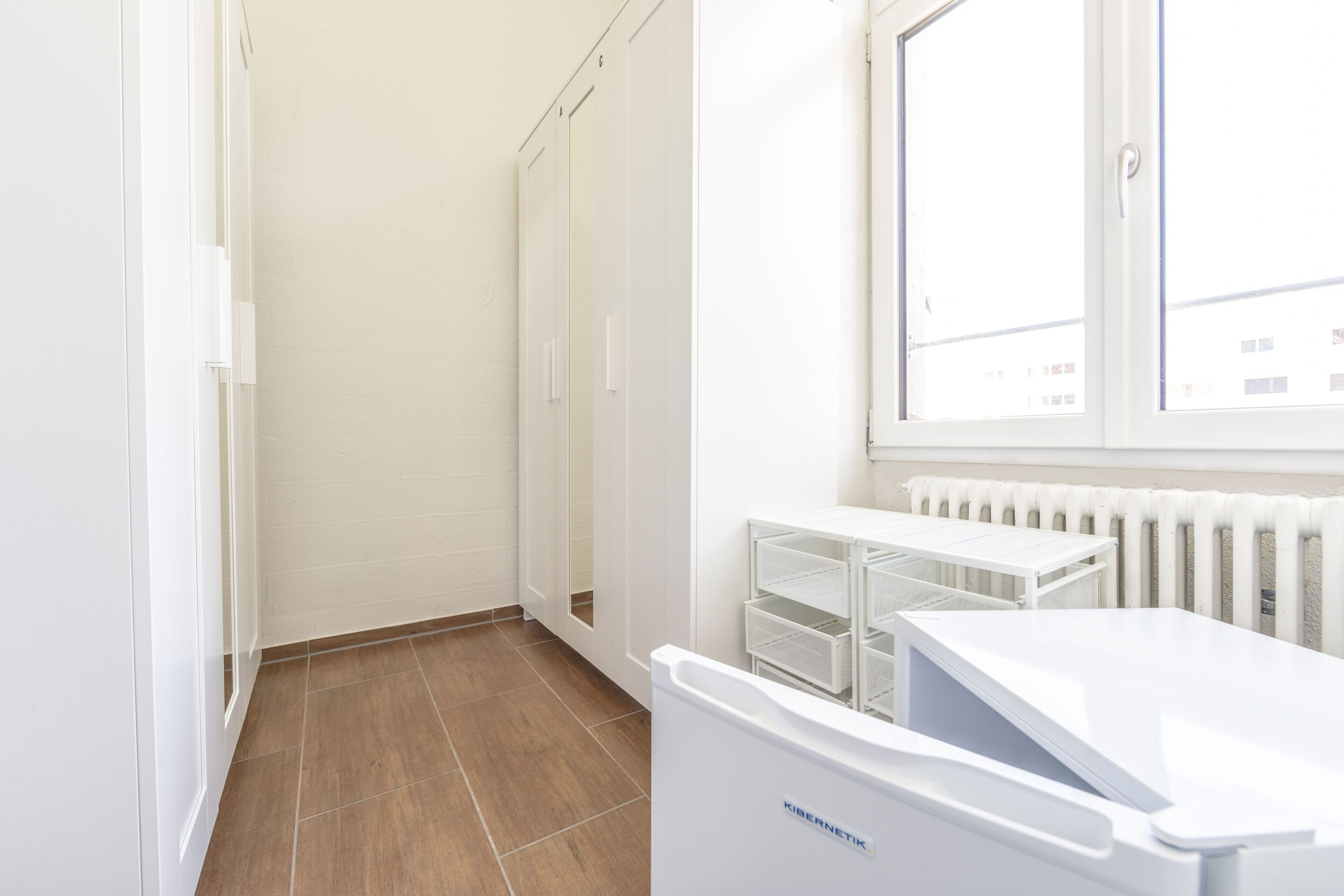 Ein sauberer, weiß gestrichener Raum mit Heizkörper, Fenster und weißem Schrank.