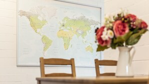 A framed world map on a white brick wall with a wooden chair and a vase of flowers on a table. - Yellow Inn (St. Gallen)