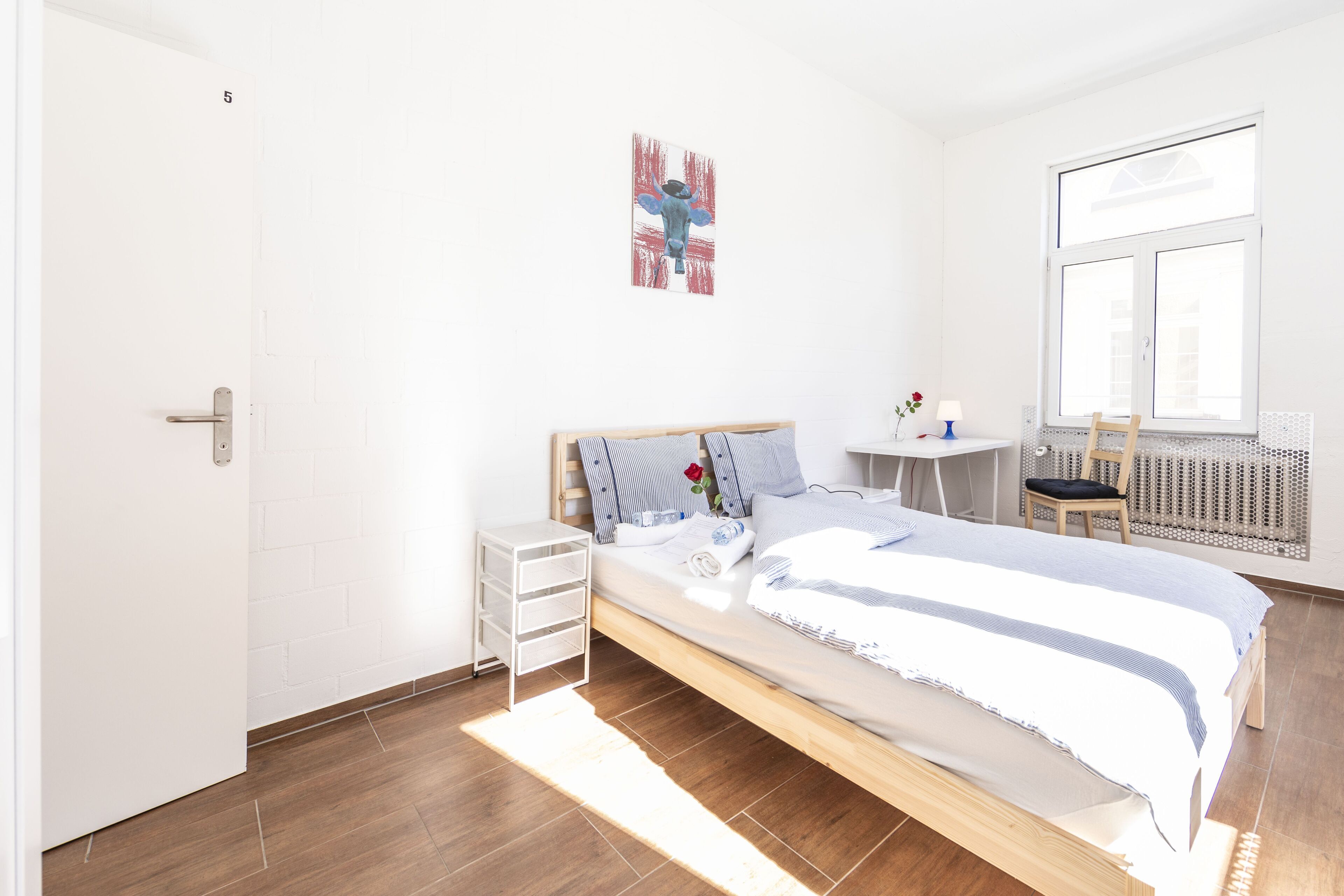 A bedroom with a wooden bed, a small bedside table, a wall-mounted artwork, a window with blinds, and a radiator.