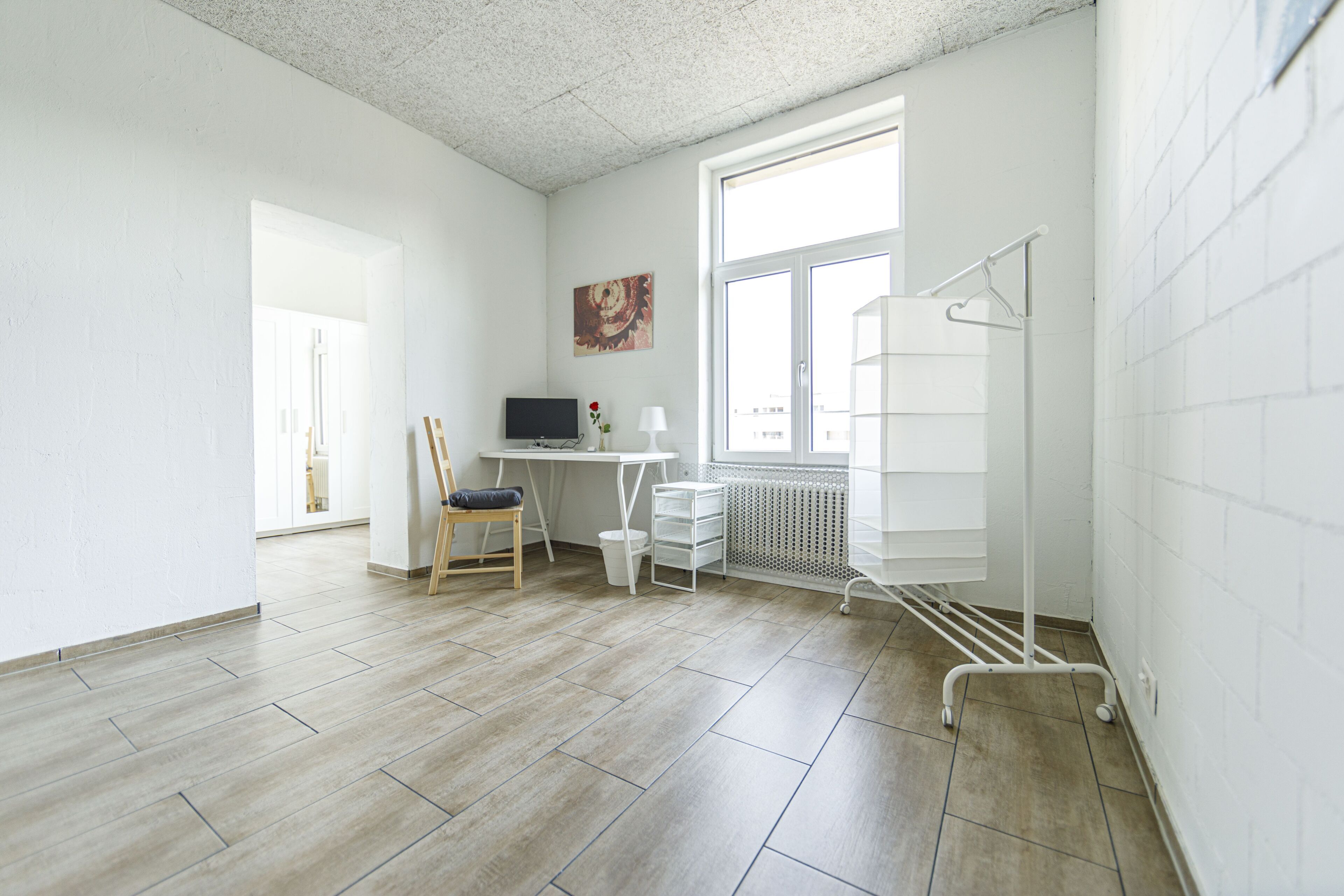 Una stanza luminosa e moderna con pavimento in legno, una scrivania con un computer, una sedia, un termosifone e una finestra con vista.