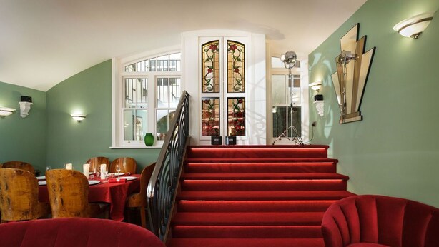 A staircase with red carpet leading to a room with green walls, a dining area with wooden chairs, and a stained glass window.