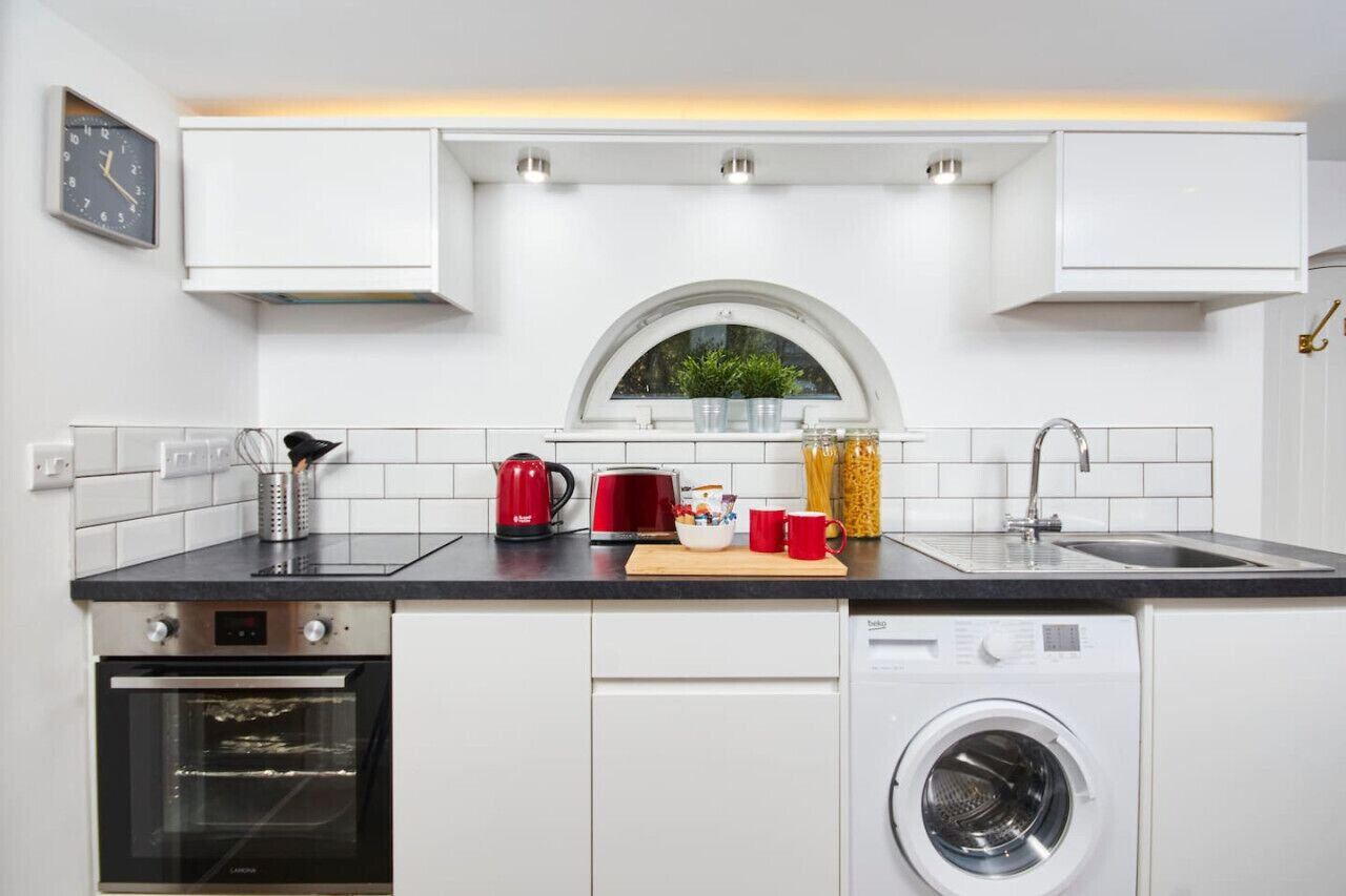 A kitchen with white cabinets, a built-in oven, a washing machine, a window, and a clock on the wall.