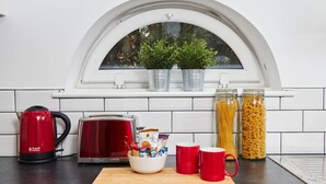 Et køkken med en hvid flisebeklædt væg, et vindue med en hvid buet top, en rød elkedel, en brødrister, en skål med snacks, to røde krus og krukker med pasta og krydderier.