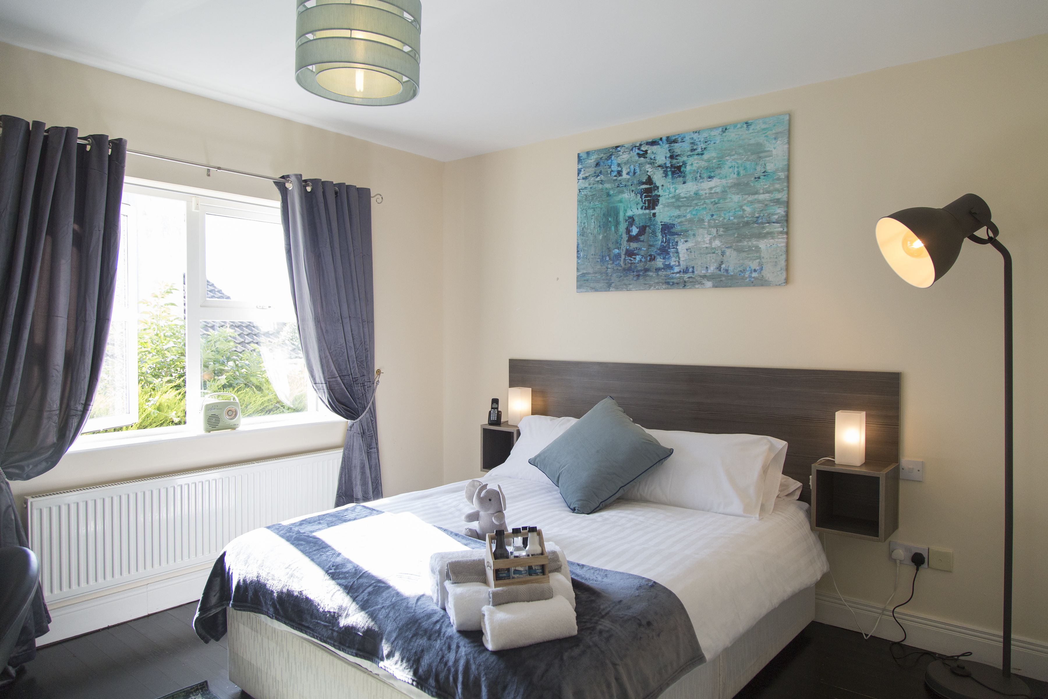 A neatly made bed with a blue pillow, a bedside table with a lamp, and a window with curtains.