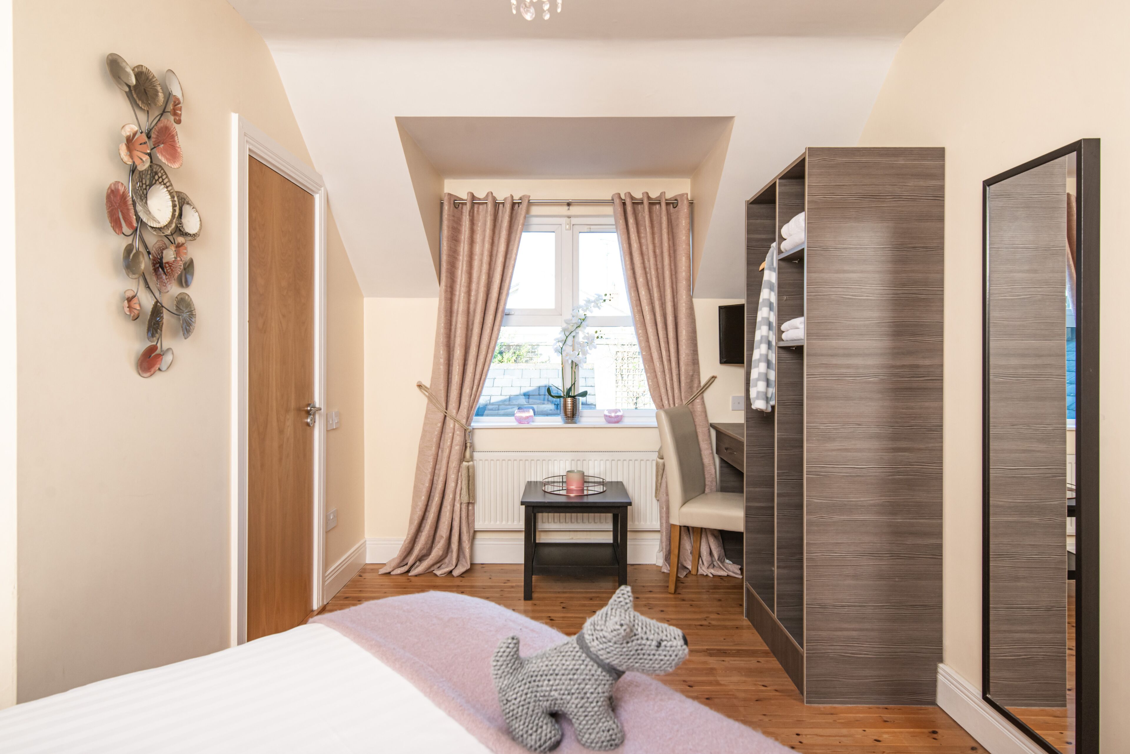 A bedroom with a bed, a window with curtains, a small table with a plant, a chair, and a wardrobe.