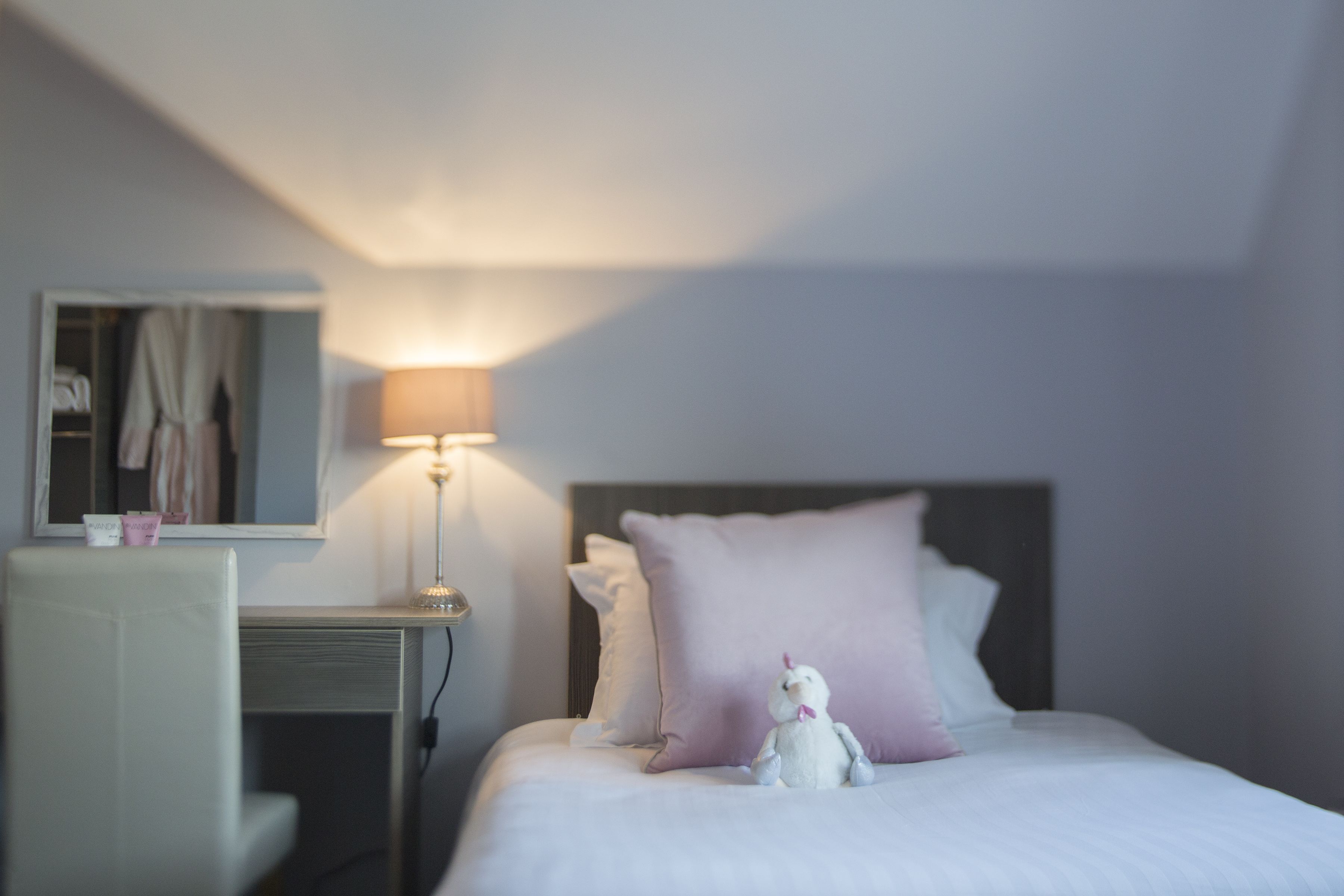 Una cama bien hecha con cabecera, lámpara de noche y un pequeño peluche sobre las almohadas.