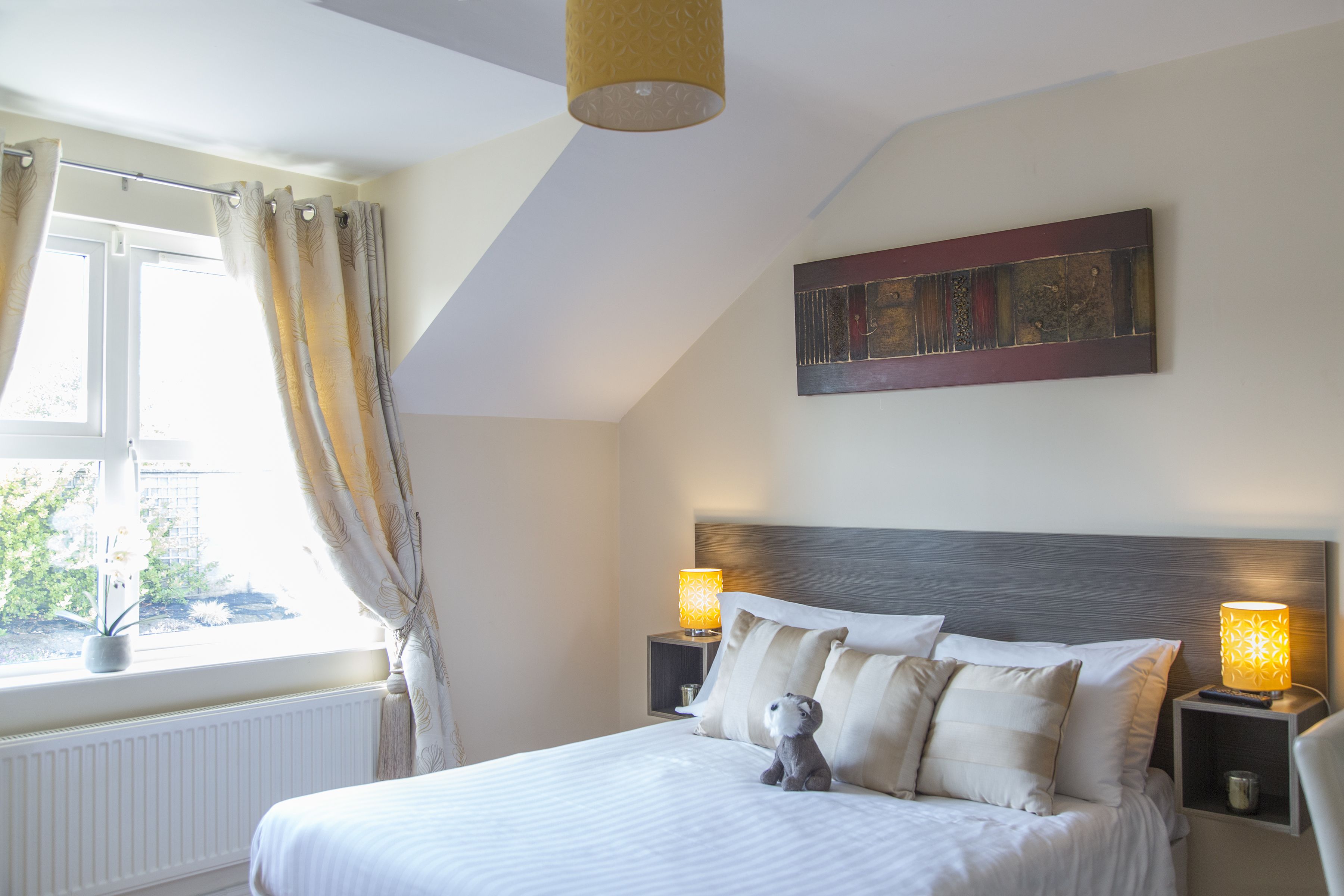 A bedroom with a bed, two bedside tables with lamps, a window with curtains, a radiator, and a wall art piece.